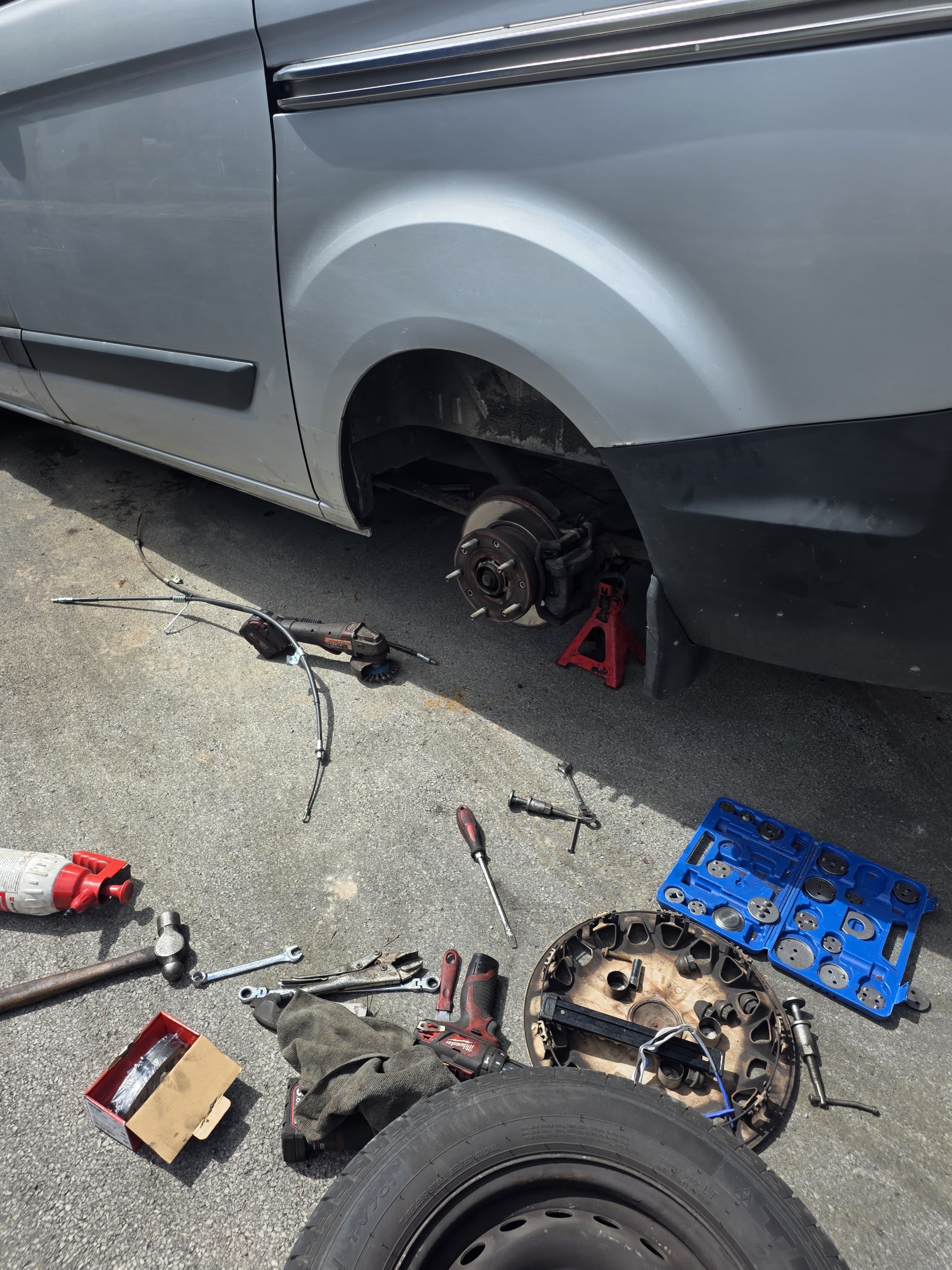 Silver van with a rear wheel off on a driveway, tools and parts on the ground